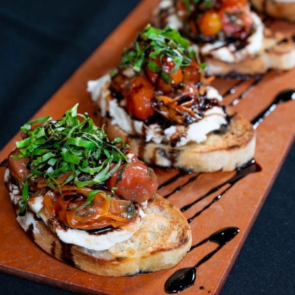 Three bruschetta appetizers with tomatoes, cheese, basil, and balsamic glaze on a wooden board.