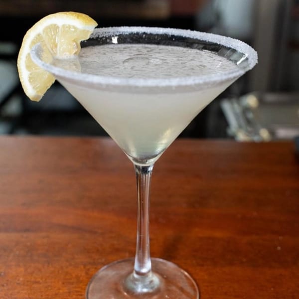 A martini glass with a sugar-rimmed edge, filled with a light-colored drink and garnished with a lemon slice.