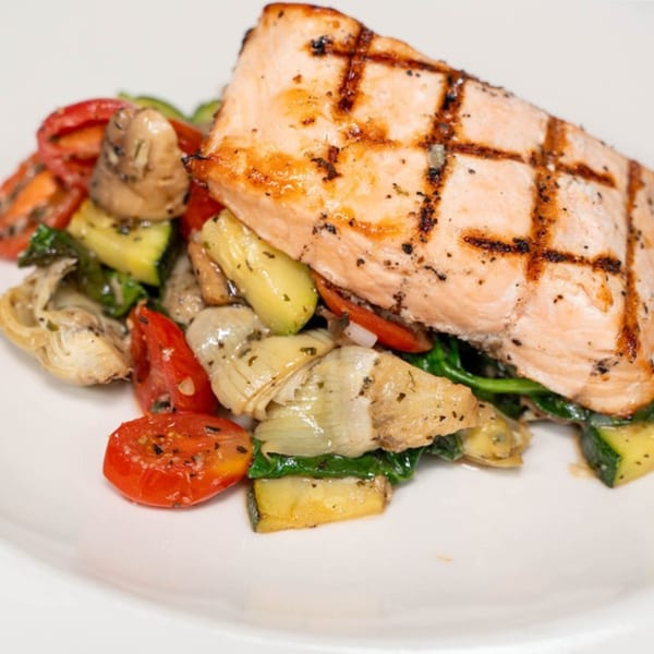 Grilled salmon fillet with charred marks, served on a bed of roasted vegetables including zucchini, tomatoes, and artichokes.